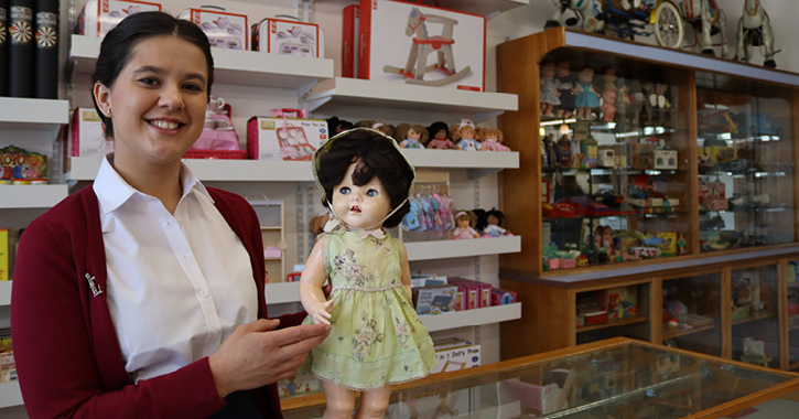 woman smiling at camera holding a vintage baby doll inside the 1950s toy shop at Beamish Museum.